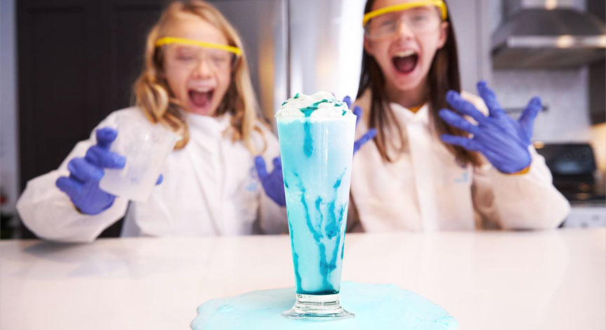 2 girls in a Mad Science lab coats,safety gloves and safety goggles making a home made milk shake that is overflowing on the 