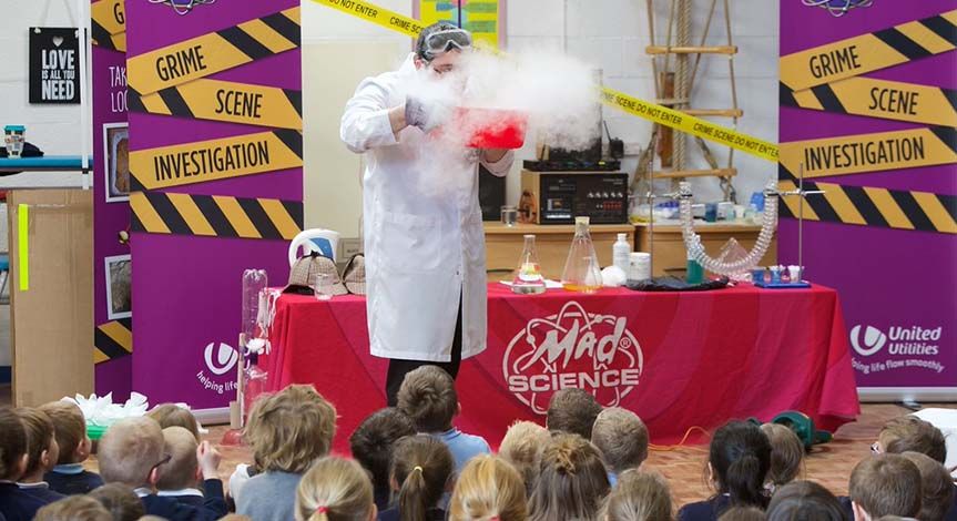 Mad scientist blowing into a red bowl with smoke coming out of it while kids watch